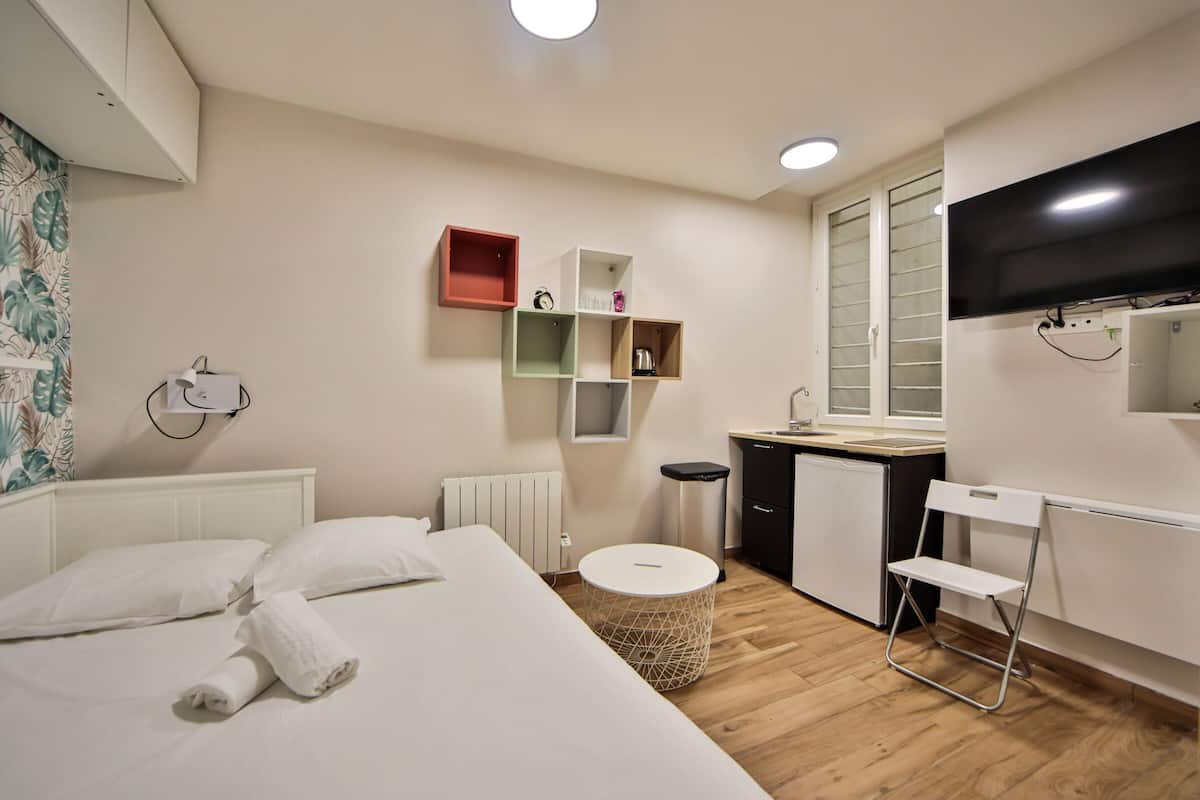 A hotel room with a bed, a small kitchenette, a TV, and a window.
