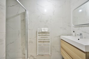 A bathroom with a glass shower enclosure, a white towel rack, a sink with a mirror, and marble tiles. - Blue Night International 49 (Paris)
