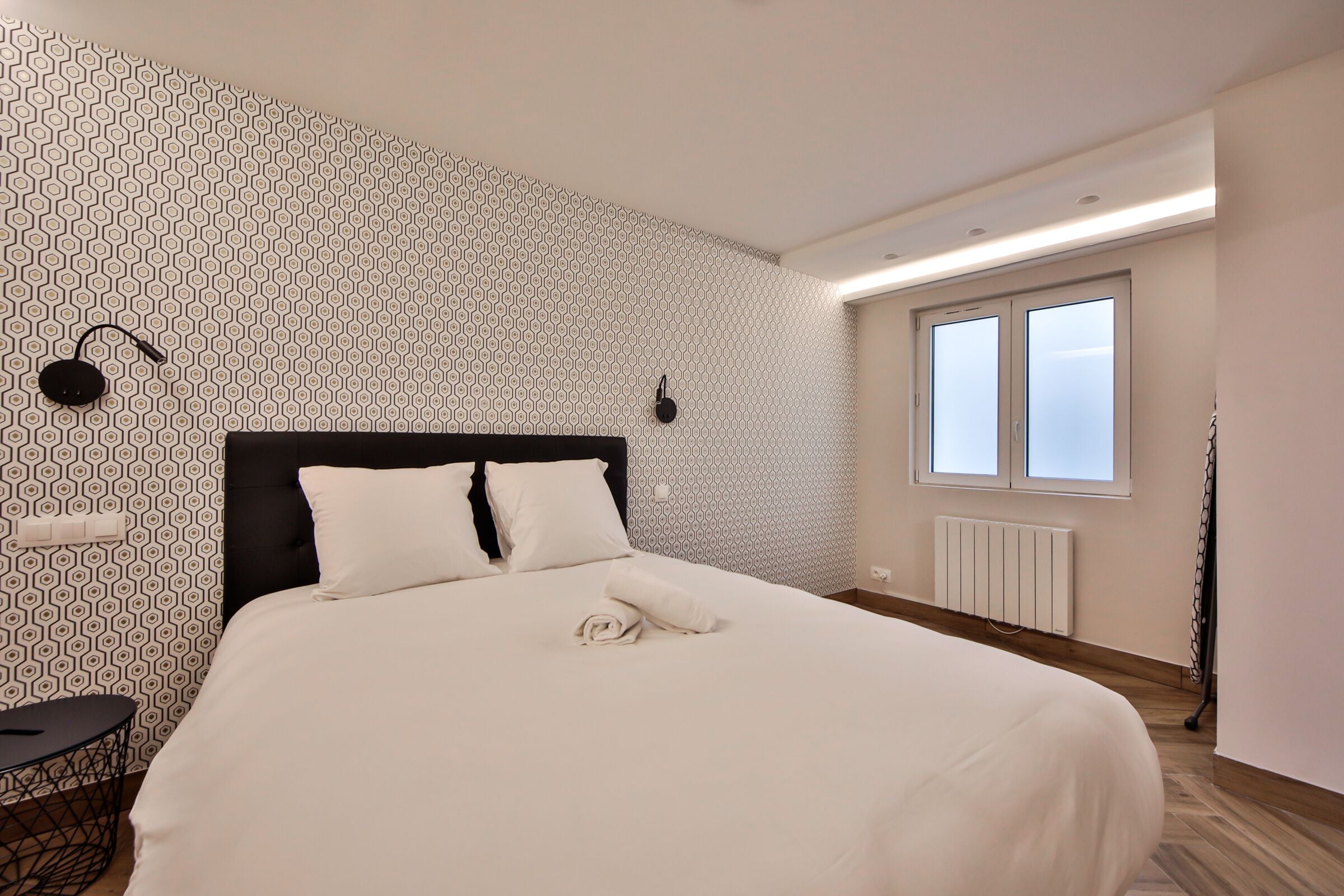 A neatly made bed with white linens, a black headboard, and a geometric patterned wall.