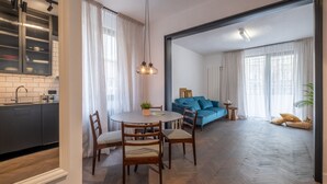 Eine moderne Küche mit dunklen Schränken und einer weißen Arbeitsplatte, ein Essbereich mit rundem Tisch und Stühlen sowie ein Wohnzimmer mit blauem Sofa und großem Fenster.