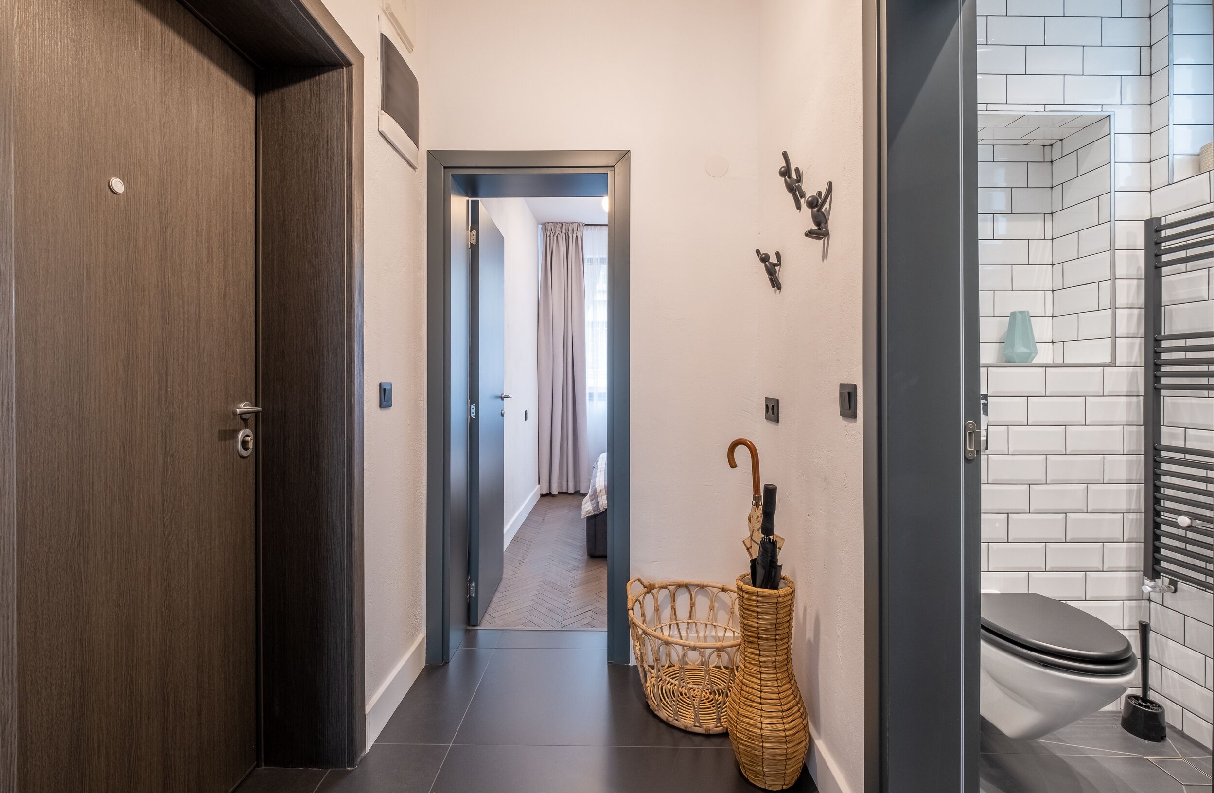 Un couloir avec une porte en bois, un panier en osier contenant un parapluie et une salle de bain carrelée.