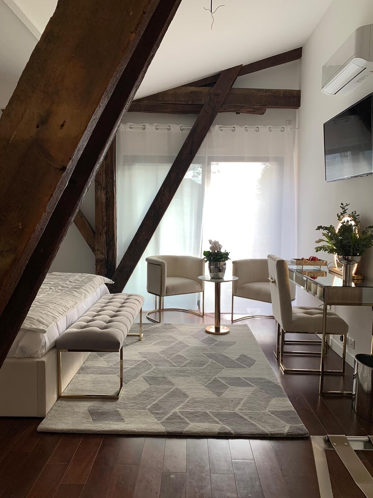 A modern loft bedroom with a sloped ceiling, a large window, a bed, a seating area with chairs, and a small table.