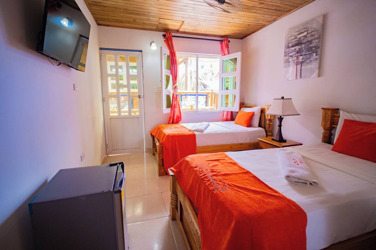 A hotel room with two beds, a television, a window with curtains, a wooden ceiling, and a painting on the wall.