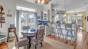 A spacious kitchen with a central island, a dining area with wicker chairs, and a large window with starfish decorations. - Beachfront Retreat | 3-Story Home | Walk to Vilano Beach | Near Nights of Lights (St. Augustine)
