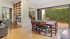 A modern living room with a wooden bookshelf, a dining table with chairs, and a view of a brick wall with greenery. - Fabulous 2BR close to the heart of East Fremantle - walk to the river (East Fremantle)