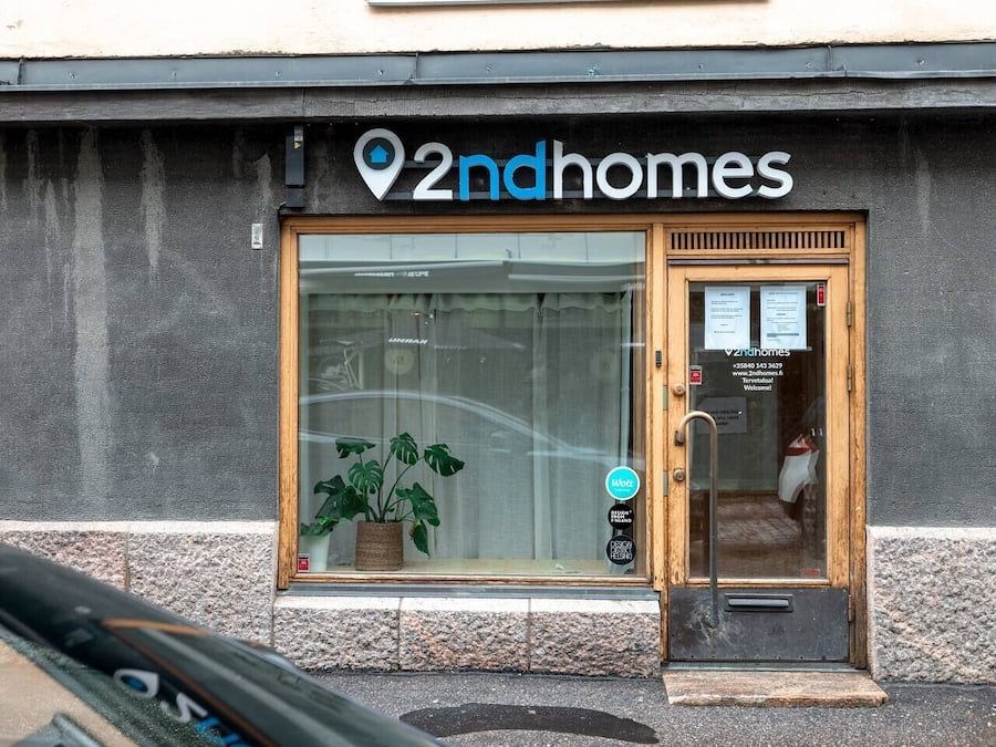 Fachada com uma grande janela de vidro, porta de madeira e uma placa com a inscrição "2ndhomes".