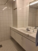 Un baño con paredes de azulejos blancos, un espejo grande, dos lavamanos y una zona de ducha.