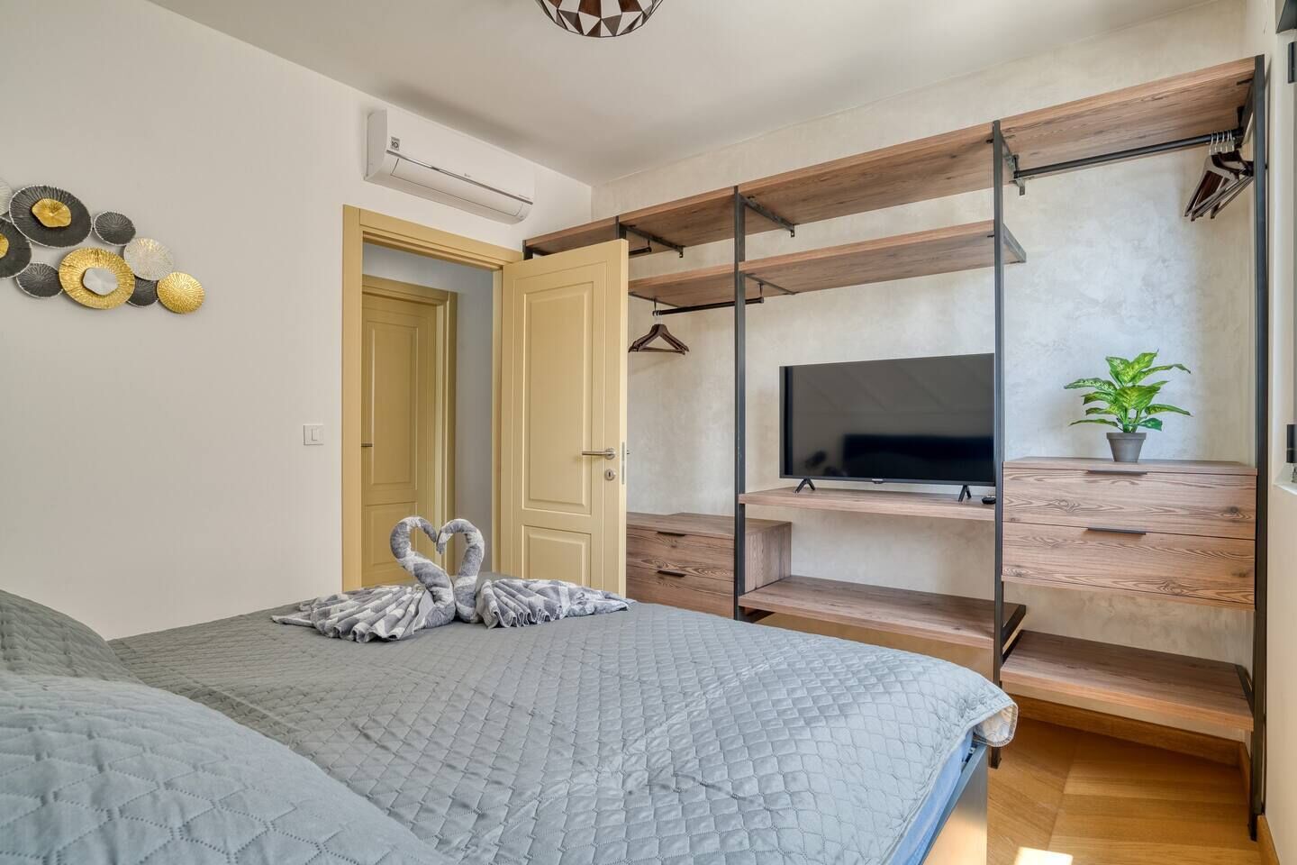 Un dormitorio con cama, televisor en un estante de madera, planta en maceta y aire acondicionado instalado en la pared.