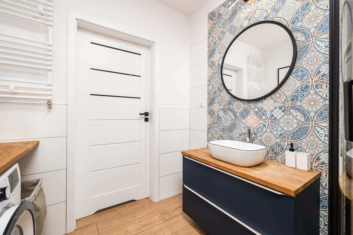Et badeværelse med en hvid dør, et rundt spejl, en vask med træbordplade og vægge med mønstret flisearbejde.
