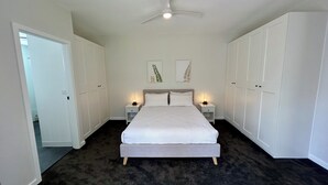 Un dormitorio con una cama, dos mesitas de noche con lámparas, un ventilador de techo y armarios blancos.