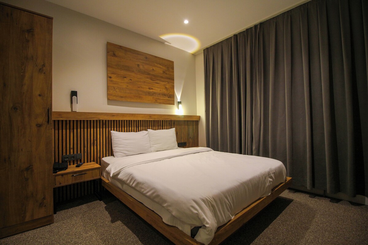 A modern hotel room with a wooden bed frame, a nightstand, a wall-mounted light, and a large window with curtains.