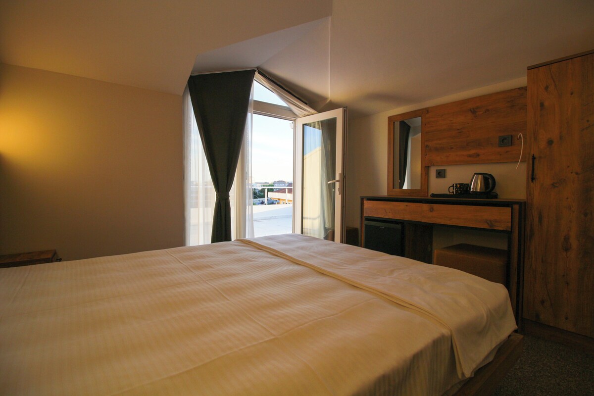 A bedroom with a large bed, a wooden desk, a mirror, and a view of the outside.