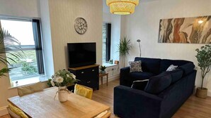 A modern living room with a dark blue sofa, a wooden dining table with chairs, a TV on a black cabinet, and a large window with a view of greenery. - Remarkable 1-bed Seaview Beach Apartment Barmouth (Barmouth)