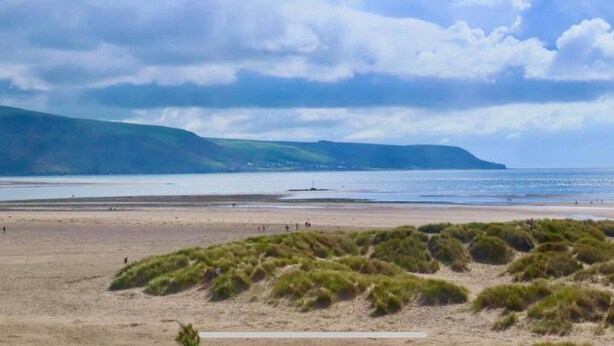 A beach with grassy dunes in the foreground, a calm sea, and distant hills under a partly cloudy sky. - Remarkable 1-bed Seaview Beach Apartment Barmouth (Barmouth)