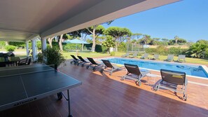 A poolside area with lounge chairs, a ping pong table, and a view of the pool and surrounding greenery.