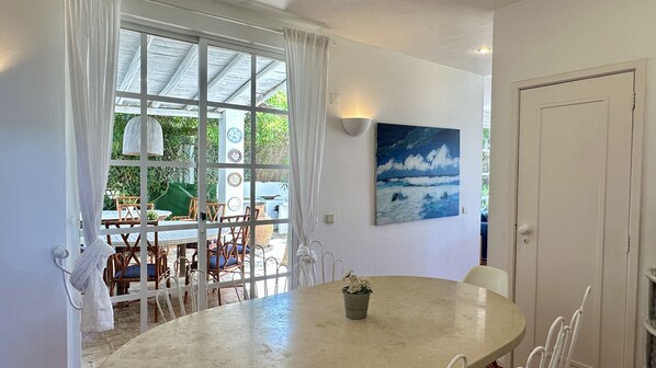 A dining area with a round table, chairs, a potted plant, and a view through a window to an outdoor seating area.