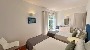 A hotel room with two beds, a chair, a desk with a mirror, and a window with curtains.