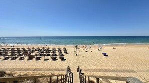 Una playa con hileras de sombrillas y tumbonas, un paseo marítimo de madera que conduce a la playa y el océano al fondo.