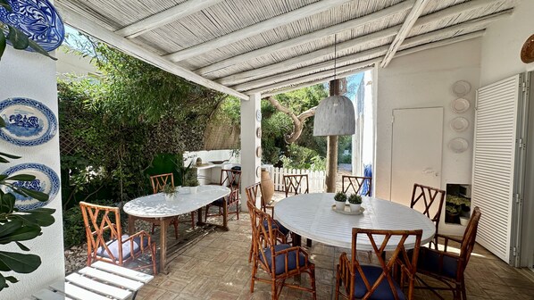 Un patio exterior con dos mesas redondas y sillas, rodeado de follaje y una pared blanca decorada con platos.