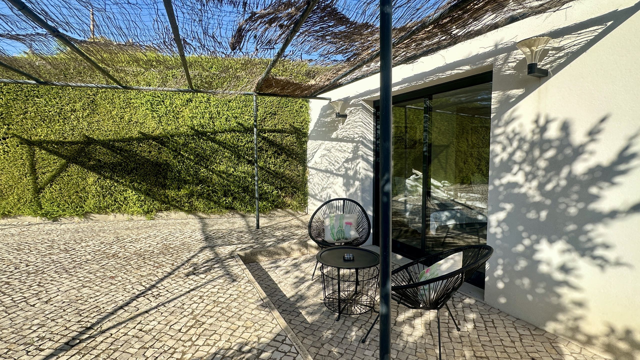 A patio with a table and chairs, a glass door, and a hedge in the background.