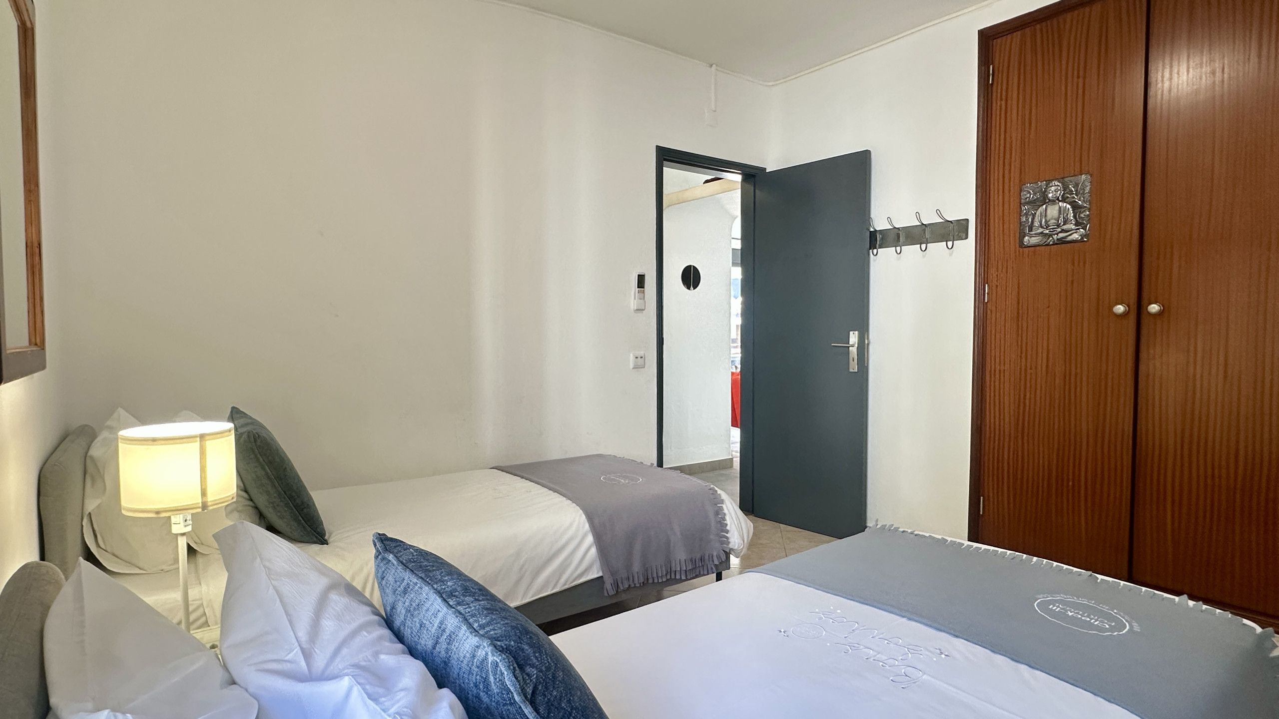 A hotel room with two beds, a wooden wardrobe, a bedside table with a lamp, and a mirror.
