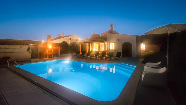 A swimming pool with lounge chairs and a table, surrounded by a building with a tiled roof and a well-lit interior. - Strip by Check-in Portugal (Albufeira)
