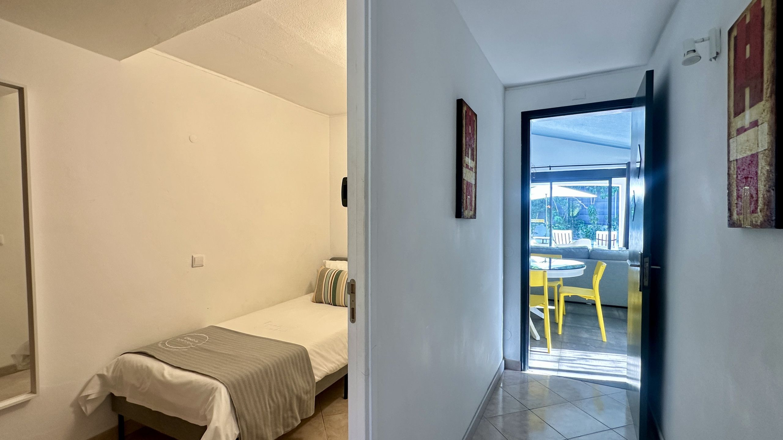 Een kleine kamer met een enkel bed, een nachtkastje en een stoel. Een deur leidt naar een balkon met een tafel en stoelen.