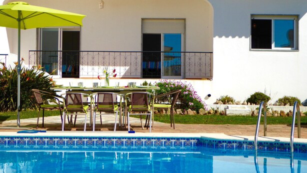Zona junto a la piscina con una mesa y sillas, una sombrilla verde y vistas a un edificio con tejado de azulejos y balcón.