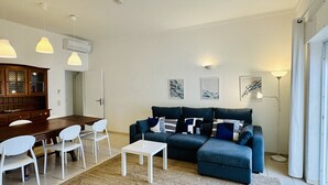 Una sala de estar con un sofá azul oscuro, sillas blancas, una mesita pequeña blanca y un aparador de madera.