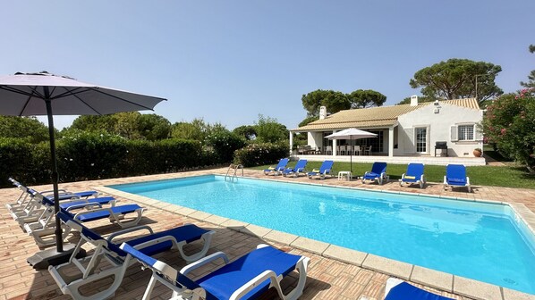 A rectangular swimming pool with blue water, surrounded by lounge chairs and umbrellas, and a building with a tiled roof in the background.