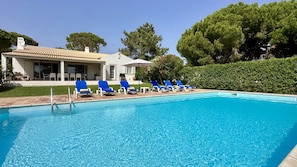 Une piscine à l’eau bleue, entourée de chaises longues et bordée d’un bâtiment dont le toit est recouvert de tuiles.