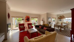 Een ruime woonkamer met een rood tapijt, een eethoek met rieten stoelen, en een keuken die door een open deur zichtbaar is.