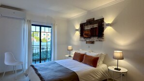 Quarto de hotel com cama, duas luminárias de cabeceira, janela com cortinas, uma cadeira e uma obra de arte pendurada na parede.