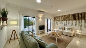 Uma sala de estar com sofá, mesa de centro e televisão, uma varanda com mobiliário de exterior e uma parede com trepadeiras.