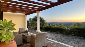 An outdoor patio with wicker furniture and a pergola, overlooking a mountainous landscape.