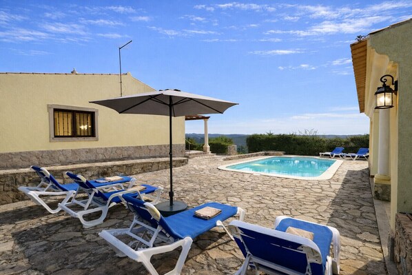 Ein Poolbereich mit Liegestühlen und Sonnenschirm, umgeben von Steinböden und Blick auf die Landschaft.