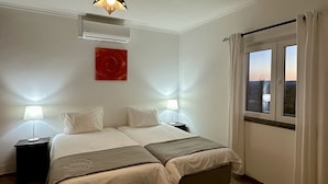 A hotel room with a double bed, two bedside tables with lamps, a red abstract painting, an air conditioning unit, and a window with curtains.