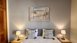 Un dormitori amb un llit, dues làmpades de nit, un quadre amb una escena de platja i una porta de fusta.