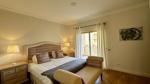 Une chambre à coucher avec un grand lit, deux tables de chevet avec des lampes, un banc et une fenêtre avec des rideaux.