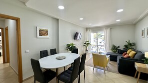 Una sala moderna con un sofá, una mesa de comedor con sillas, un televisor montado en la pared y un balcón con vistas.
