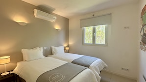 Quarto com duas camas, janela com vista, ar condicionado e luminária de cabeceira.