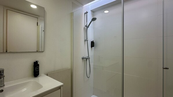 A bathroom with a white sink, a mirror, a shower with a handheld showerhead, and a handheld soap dispenser.