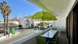 A balcony with a table and chairs, a green umbrella, and a view of the street with buildings and palm trees. - Medronheira by Check-in Portugal (Albufeira)