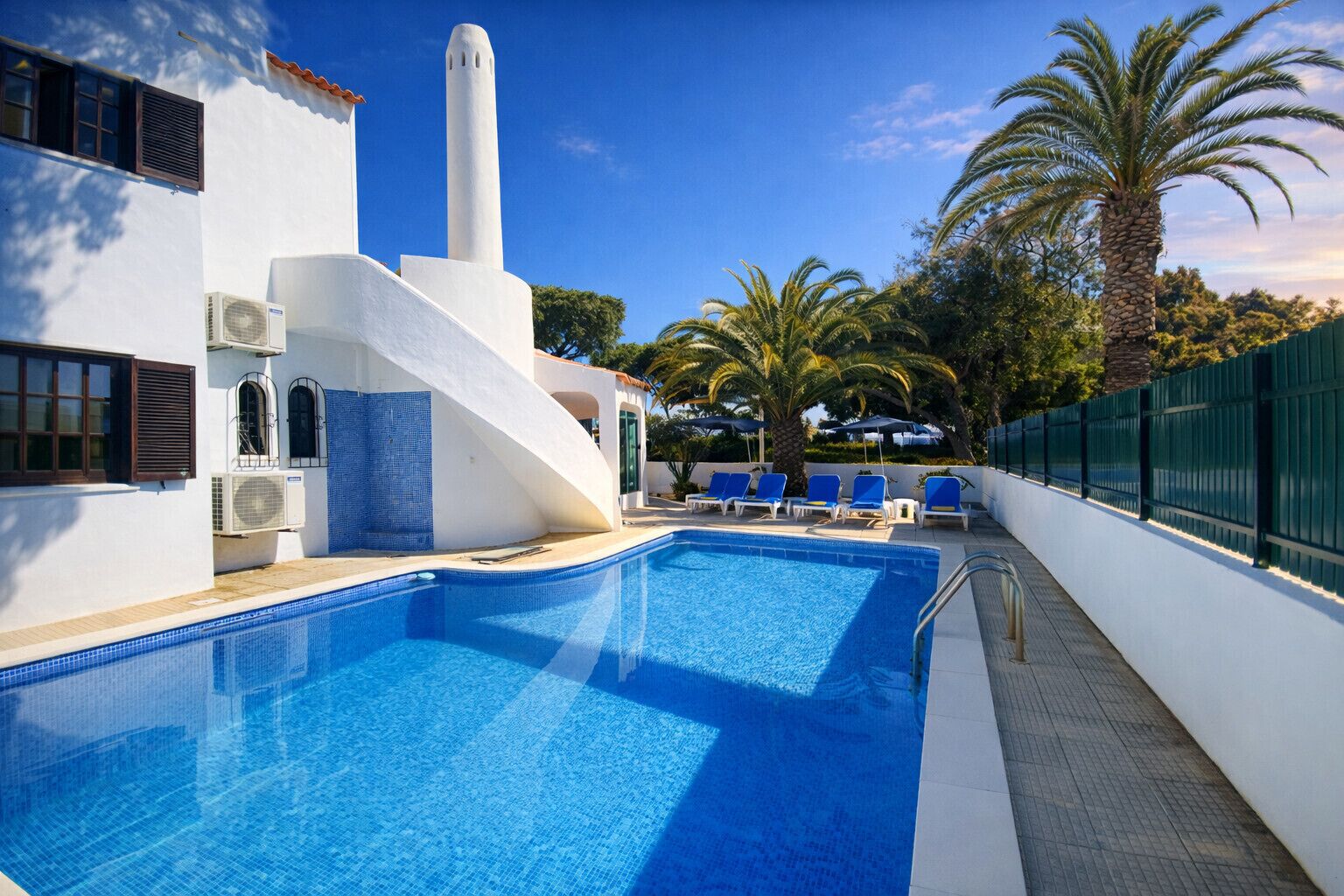 A swimming pool with a sun terrace, palm trees, and a white building with a tall chimney.