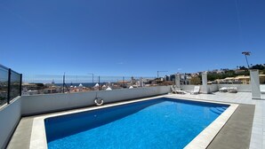 A rooftop swimming pool with clear blue water, surrounded by a white tiled deck and a metal fence.