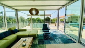 En moderne stue med sofa, sofabord og stol. Stuen har store glasvinduer og -døre med udsigt til et poolområde og grønne omgivelser.