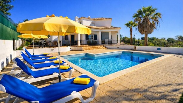 A pool area with blue lounge chairs and yellow umbrellas, a clear blue sky, and a white building with a tiled roof. - Abby by Check-in Portugal (Albufeira)