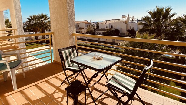 A balcony with a table and chairs, a pool, and a view of buildings and palm trees.