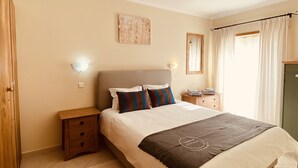 A bedroom with a bed, bedside tables, a nightstand, a wall-mounted light, a mirror, and a window with curtains.