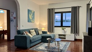 Ein Wohnzimmer mit einem Türkis-sofa, einem kleinen runden Tisch, einem großen Fenster mit Vorhängen, einem Gemälde an der Wand und einem Teppich auf dem Boden.